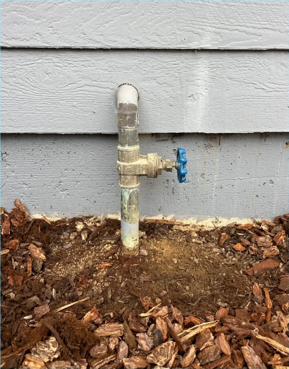 Old leaking main water shutoff valve with aging galvanized and copper piping installed beside a residential home foundation prior to replacement.