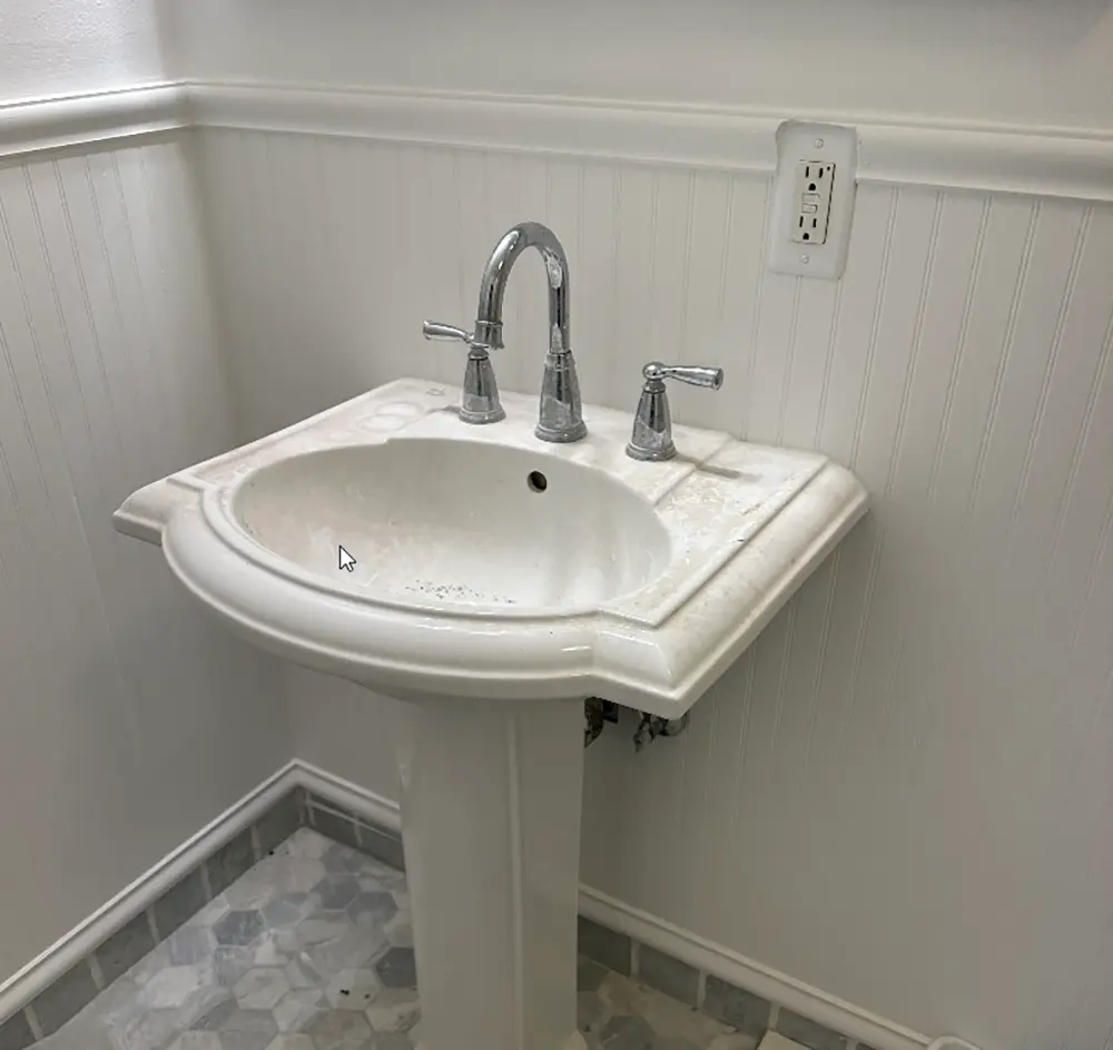Existing widespread faucet installed on a bathroom pedestal sink before replacement, showing the original faucet and drain assembly prior to plumbing upgrade.