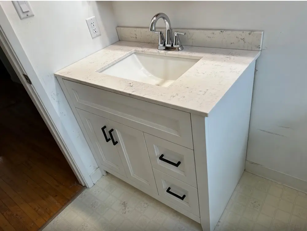 New bathroom vanity replacement featuring a quartz countertop, undermount sink, decorative backsplash, soft-close cabinet doors, and fully sealed installation viewed from an angled perspective.