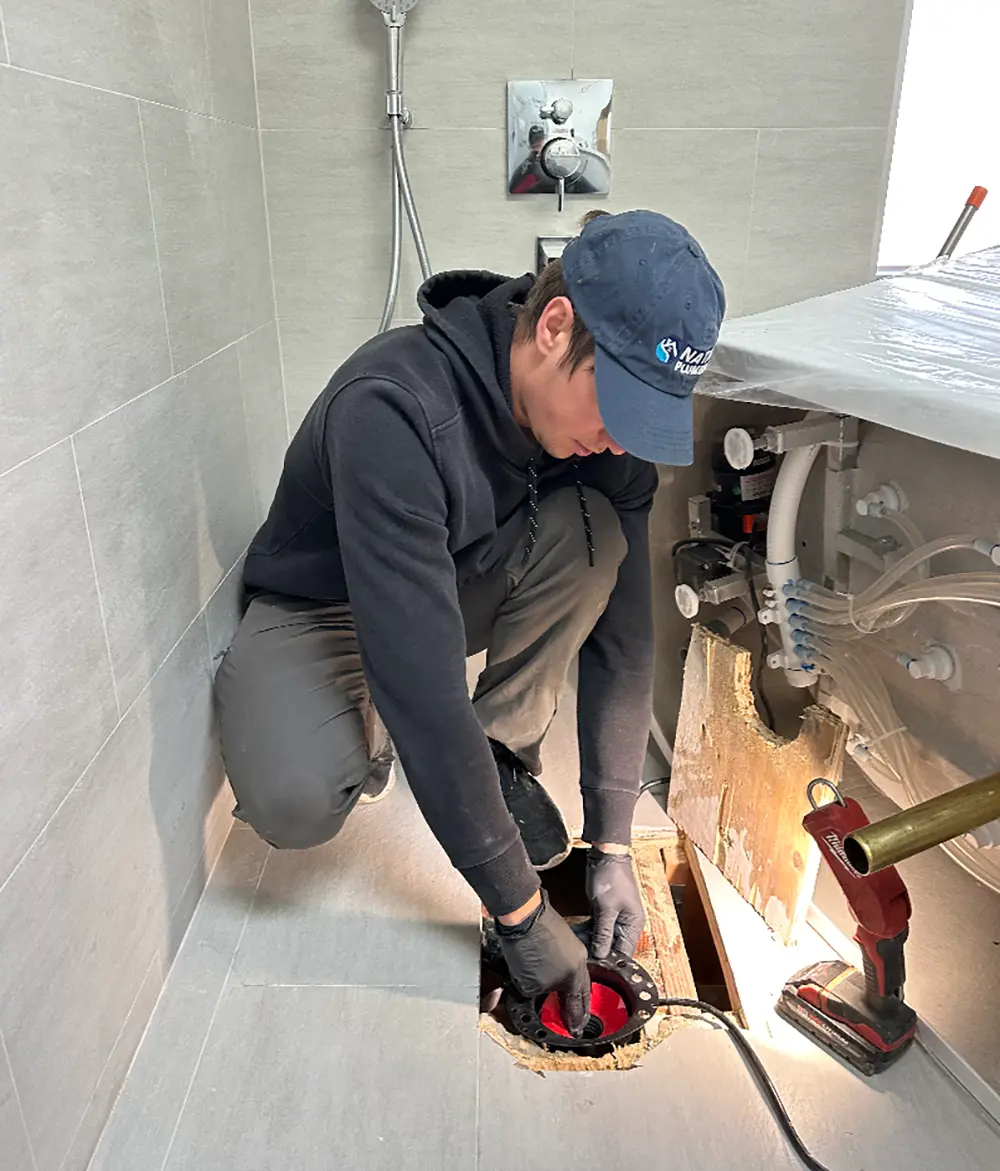 Technician installing the drain assembly and leveling a freestanding bathtub during professional bathroom plumbing installation prior to final placement and sealing.