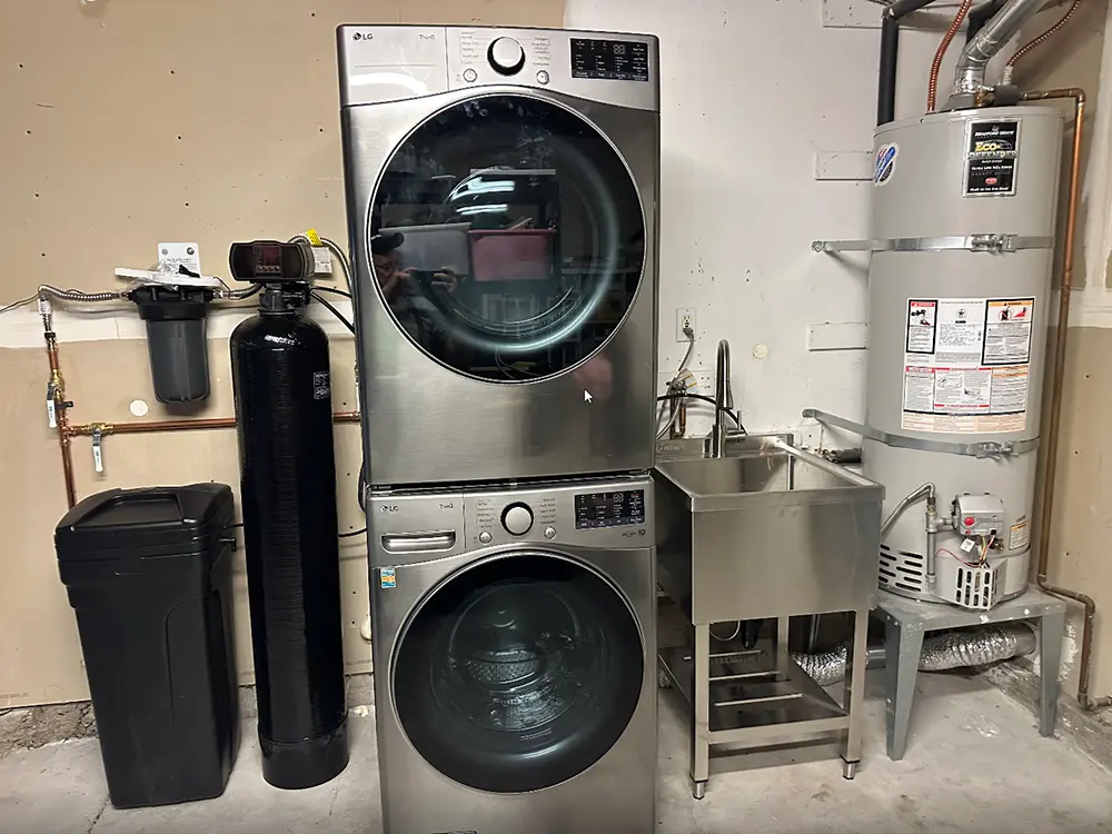 Garage laundry room featuring stacked washer and dryer installation, new utility sink, water softener system, and upgraded plumbing for a space-saving laundry renovation.