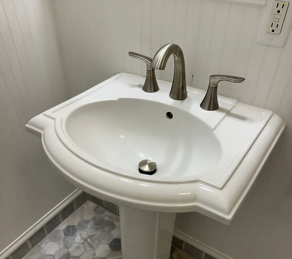 New widespread bathroom faucet professionally installed on a pedestal sink with reconnected drain assembly, silicone sealing, and leak-free plumbing connections.
