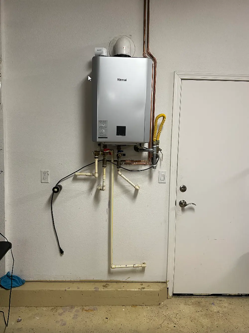 Existing tankless water heater installed on a garage wall before replacement, featuring the original plumbing, venting, and gas line connections.